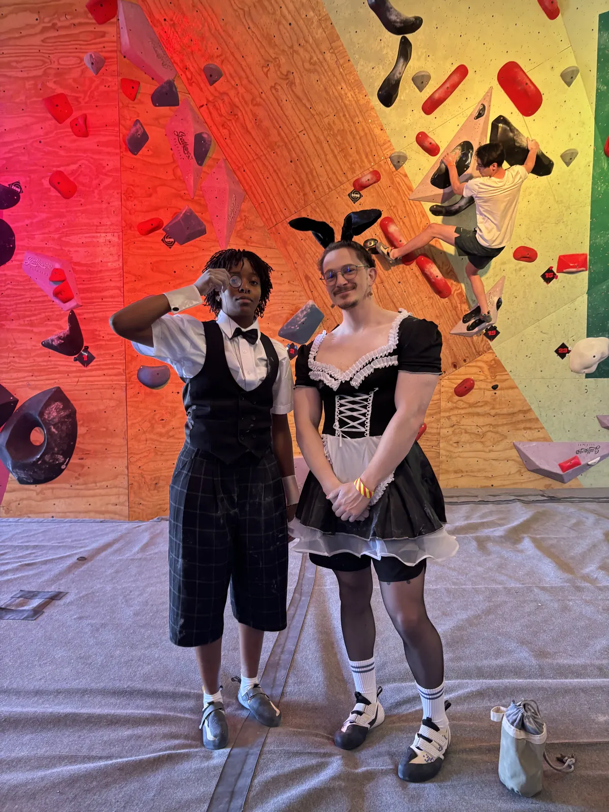 Cedric in a maid outfit and their tjom dressed as a butler, standing in front of the climbing wall